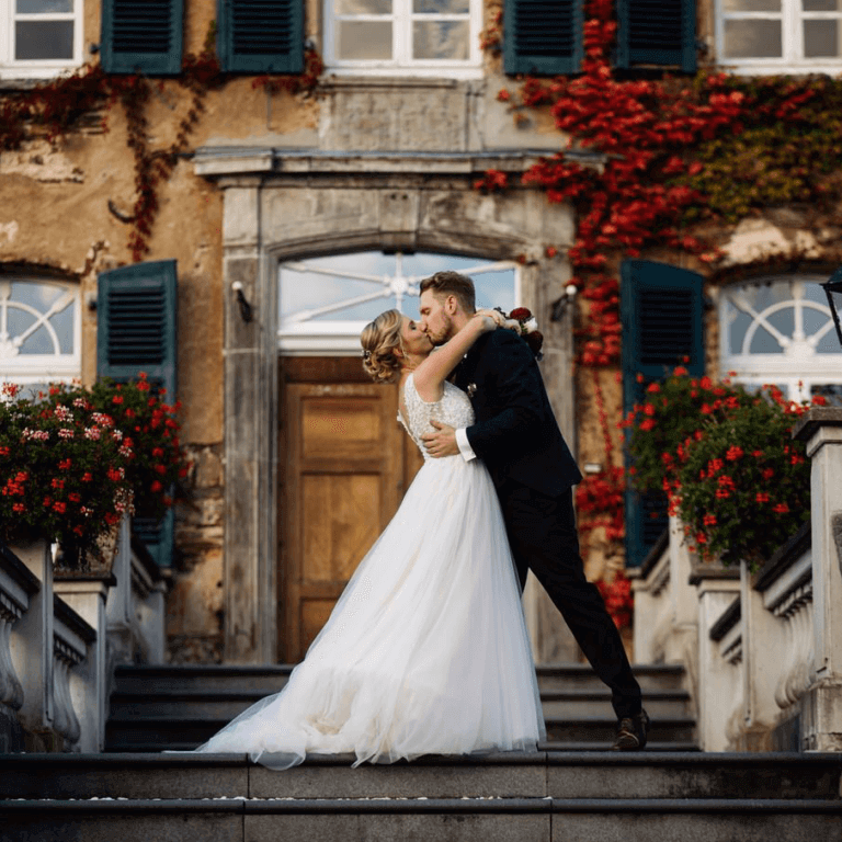 DJ Hochzeit Hochzeits-DJ Eine Braut und ein Bräutigam teilen sich bei ihrer Hochzeit in Köln einen romantischen Kuss auf den Stufen eines prächtigen Herrenhauses.